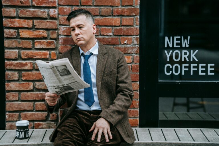 A professional man in formal wear reads a newspaper beside a coffee shop window.